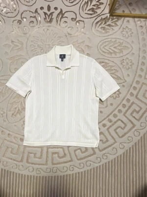 Vit ribbad pikétopp  - Vit pikétopp  med korta ärmar och klassisk krage. Toppen har diskret ribbad struktur och en stilren, enkel design som passar till många olika outfits. Perfekt för dig som gillar en clean och fräsch look.