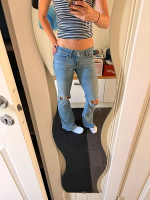 Ljusblå bootcut jeans med hål - Säljer ett par ljusblå bootcut jeans med slitna hål på båda knäna. Jeansen har klassisk femficksmodell och normal midja. Perfekta för en avslappnad och trendig look. Materialet är denim i bomull.