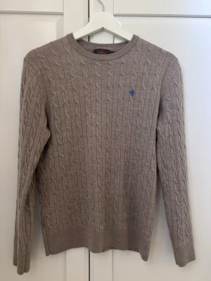 Morris tröja kan gå ner pris  - Kabelstickad tröja i beige med rund halsringning och lång ärm i str M. Tröjan har ett diskret blått broderat emblem på bröstet och ribbade muddar vid ärmslut och nederkant. Perfekt för dig som gillar klassisk och stilren design.
