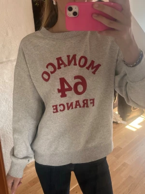 Grå sweatshirt med röd text - Säljer en grå sweatshirt med röd text framtill där det står 'MONACO 64 FRANCE'. Tröjan har rund halsringning, långa ärmar och ribbade muddar vid ärmslut och nederkant. Perfekt för en avslappnad och sportig stil.