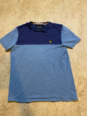 Tvåfärgad blå t-shirt från Lyle & Scott - Snygg t-shirt från Lyle & Scott i två nyanser av blått. Övre delen är mörkblå och resten ljusblå, med den klassiska gula loggan på bröstet. T-shirten har rund hals och korta ärmar, perfekt för en avslappnad stil.