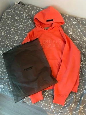 Korallfärgad Essentials hoodie - Snygg korallfärgad hoodie från Essentials Fear of God med broderad logga på bröstet och patch på huvan. Klassisk känguruficka framtill och ribbade muddar. Perfekt för en avslappnad och trendig streetwear-look.