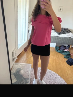 Rosa topp med volangdetaljer - Säljer en söt rosa topp med volangdetaljer över bröstet och puffiga korta ärmar. Toppen har en rak passform och är gjord i ett mjukt, tunt tyg som känns skönt mot huden. Perfekt för dig som vill ha en färgglad och trendig look.