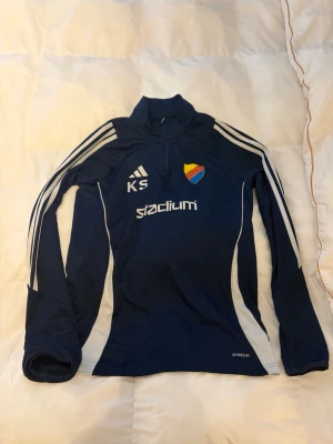 Blå träningströja Djurgården Adidas - Mörkblå långärmad träningströja från Adidas med Djurgårdens IF klubbmärke på bröstet, vita detaljer och dragkedja vid halsen. Tre vita ränder längs ärmarna och texten 'stadium' framtill. Tillverkad i funktionsmaterial. Har ett pyttelitet hål som knappt syns. ABS! Pris går att diskutera!