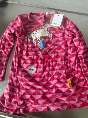 Rosa Pippi Långärmad Klänning - Söt långärmad klänning från Polarn O. Pyret med rosa och röda geometriska mönster. Framtill finns ett tryck av Pippi Långstrump och Lilla Gubben samt ficka med Pippi-märke. Perfekt för dig som älskar färg och lekfull stil.