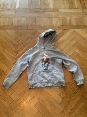 Grå Ralfh lauren hoodie  - Säljer en grå hoodie från Ralfh lauren med huva och ett tryck av en björn. Hoodien har ribbade muddar vid ärmslut och nederkant. Perfekt för dig som gillar streetwear och vill ha något bekvämt och stilrent med en rolig twist. Nypris är runt 1700kr mitt pris är 399kr. Str är 164 cm typ. Skriv dm för frågor.