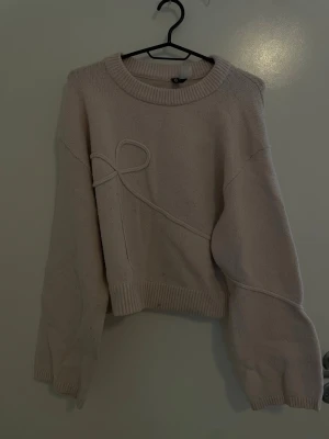 Ljusbeige stickad tröja H&M Divided - Supersöt ljusbeige stickad tröja från H&M Divided med ribbade muddar och en unik dekorativ snodd i form av en rosett på framsidan. Tröjan har rund halsringning och avslappnad passform, perfekt för lager på lager under kyliga dagar.