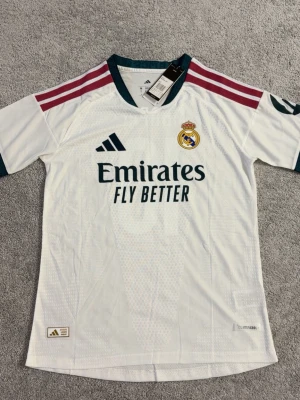 Real Madrid matchtröja vit Adidas - Säljer en officiell Real Madrid matchtröja från Adidas i vitt med mörkgröna detaljer och röda axelränder. Tröjan har klubbmärke på bröstet, sponsortryck och är tillverkad i ett lätt, ventilerande funktionsmaterial. Perfekt för fotbollsträning eller att bära till vardags.