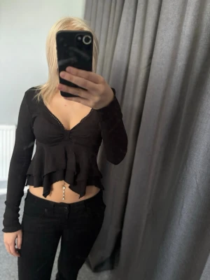 Svart croppad volangtop med v-ringning - Säljer en svart croppad topp med långa ärmar och v-ringning. Toppen har volangdetaljer nedtill och en lätt ribbad struktur i materialet. Perfekt för dig som gillar trendiga och feminina plagg med snygga detaljer.