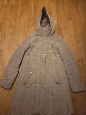 Lång beige dunjacka med huva - Snygg lång beige dunjacka med huva och tryckknappar framtill. Jackan har två snedställda bröstfickor med dragkedja och två fickor med lock och knapp. Quiltad design och figurnära passform, perfekt för kalla vinterdagar.