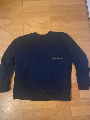 Mörkblå sweatshirt från Arigato - Snygg mörkblå sweatshirt från Arigato med gul text på bröstet. Tröjan har rund halsringning, långa ärmar och ribbade muddar vid ärmslut och nederkant. Perfekt för en avslappnad och stilren look.