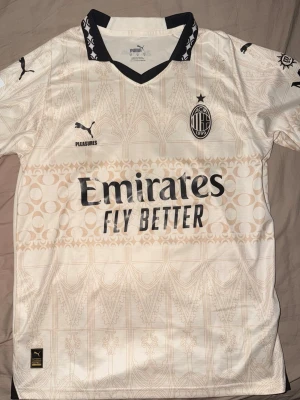 AC Milan bortatröja Puma Rafa Leão - Snygg AC Milan bortatröja i beige med svarta detaljer och grafiskt mönster. Puma-logga, klubbmärke och sponsortryck framtill. På ryggen står det 'Rafa Leão 10' och 'wefox'. Tröjan har korta ärmar och klassisk krage med svart kant.