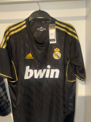 Real Madrid bortatröja Ronaldo #7 - Snygg svart Real Madrid bortatröja från Adidas med gula detaljer och tryck. Tröjan har klubbmärke, Adidas-logga och sponsortryck fram samt Ronaldo och nummer 7 i guld på ryggen. 