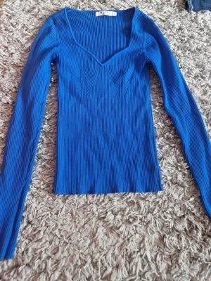 Kornblå ribbad stickad topp - Snygg kornblå stickad topp med ribbad struktur och lång ärm. Djup v-ringning och figurnära passform som ger en modern look. Perfekt att styla med jeans eller kjol för en trendig outfit.