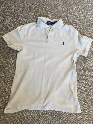 Vit pikétröja från Polo Ralph Lauren - Klassisk vit pikétröja från Polo Ralph Lauren med korta ärmar och blå broderad logga på bröstet. Tröjan har krage och knäppning med två knappar framtill. Perfekt till en avslappnad men ändå stilren look. Passar 164-170 slim fit