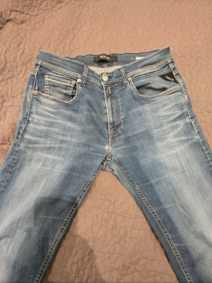 Blå Replay jeans med slitningar - Säljer ett par snygga blå jeans från Replay med klassisk femficksmodell och ljusa slitningar framtill. Jeansen har läderpatch med Replay-logga bak och kontrastsömmar. Perfekta för en avslappnad och trendig look.