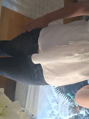 Vit broderad linnetopp med knyt - Söt vit linnetopp med tunna axelband och blå broderade detaljer längs v-ringningen. Toppen har knyt i midjan och är lätt utsvängd nedtill. Perfekt för varma dagar och ger en fräsch sommarkänsla.