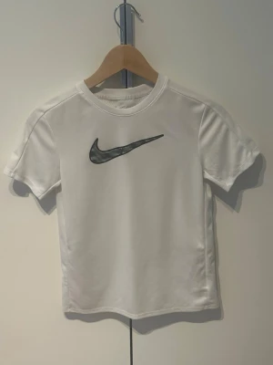 Vit Nike t-shirt med logga - Snygg vit t-shirt från Nike med svart logga på bröstet. T-shirten har rund halsringning och korta ärmar. Tillverkad i ett lätt och andningsbart material som passar perfekt till träning eller vardagsstil.