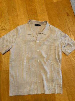 Beige stickad skjorta från J.Lindeberg - Snygg beige skjorta från J.Lindeberg i stickad kvalitet med korta ärmar och knappar framtill. Skjortan har ett diskret ribbat mönster och klassisk krage, perfekt för en clean och modern look.
