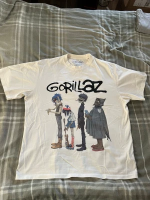 Vit Gorillaz t-shirt - Säljer en vit t-shirt med stort Gorillaz-tryck framtill. Motivet visar de tecknade bandmedlemmarna i färg och loggan ovanför. T-shirten har korta ärmar och rund hals. Perfekt för dig som gillar bandmerch och vill sticka ut med en cool design.