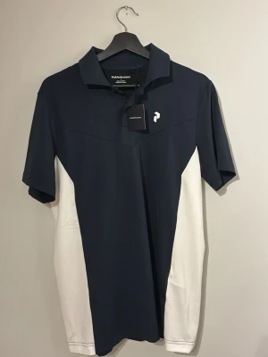 Helt ny och oanvänd med prislapp kvar. Mörkblå och vit pikétröja Peak Performance Golf  - Snygg mörkblå pikétröja från Peak Performance med vita paneler på sidorna. Tröjan har klassisk krage, korta ärmar och diskret logga på bröstet. Tillverkad i ett lätt och funktionellt material som passar perfekt till sport och träning.