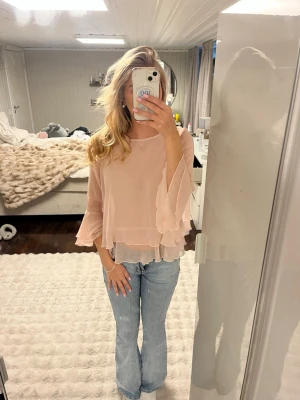 Ljusrosa volangblus  - Söt ljusrosa blus i transparent, lätt material med volangdetaljer och vida, långa ärmar. Blusen har en lös passform och rund halsringning, perfekt för en trendig och feminin look.