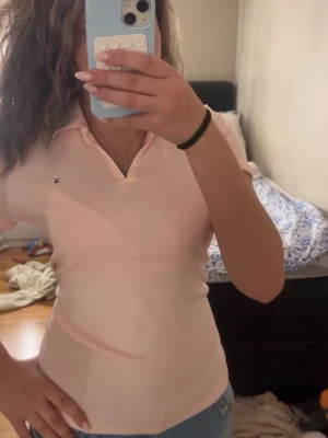 Ljusrosa pikétopp från Tommy Hilfiger - Säljer en ljusrosa pikétopp från Tommy Hilfiger med liten logga på bröstet. Toppen har krage, korta ärmar och en v-ringad öppning framtill. Perfekt till jeans eller kjol för en clean och fräsch look.