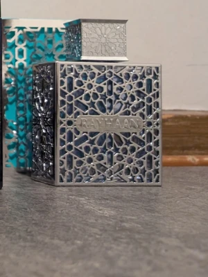 Sälj / byte Rayhaan Aquatica  - Unik parfymflaska med namnet Rayhaan, täckt av ett detaljerat silvrigt metallmönster i geometrisk stil. Flaskan är fyrkantig och har en matchande dekorativ kork. Bakgrunden visar en turkos och vit förpackning med liknande mönster.