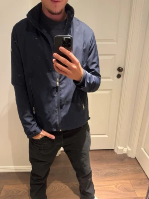 Mörkblå Moncler jacka med dragkedjor - Snygg mörkblå jacka från Moncler med ståkrage och två vertikala dragkedjefickor framtill. Jackan har en diskret Moncler-logga på vänster ärm och är tillverkad i ett slätt, lätt material. Perfekt för dig som vill ha en stilren och exklusiv look.