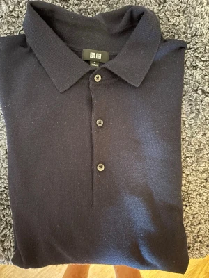Mörkblå stickad polotopp Uniqlo - Snygg mörkblå stickad polotopp från Uniqlo med klassisk krage och tre knappar framtill. Perfekt till jeans eller kjol för en clean och stilren look. 100% merinoull.