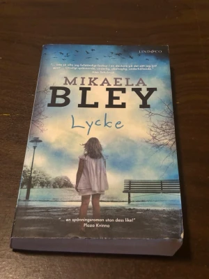 Lycke av Mikaela Bley pocket - Spännande pocketbok av Mikaela Bley med titeln 'Lycke'. Omslaget har en blå ton med en flicka som står vid en parkbänk och träd i bakgrunden. Boken ges ut av Lind & Co och är en kriminalroman som utspelar sig i Stockholm.