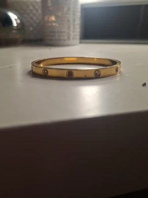 Guldfärgat armband med stenar - Snyggt guldfärgat armband i metall med infällda runda stenar runt om. Armbandet har en stilren och modern design med slät yta och diskreta detaljer. Perfekt accessoar för att lyfta din outfit.