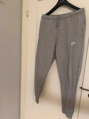 Gråa mjukisbyxor från Nike - Snygga gråa mjukisbyxor från Nike med dragsko i midjan och ribbade muddar vid bensluten. Byxorna har två sidofickor och en broderad Nike-logga på vänster ben. Perfekta för chill dagar eller träning.