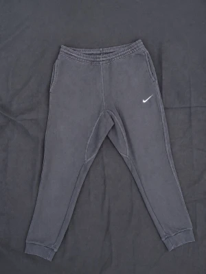 Mörkgrå mjukisbyxor från Nike - Säljer ett par mörkgrå mjukisbyxor från Nike med klassisk vit logga på vänster ben. Byxorna har resår i midjan, muddar vid bensluten och två sidofickor. Perfekta för chill dagar eller träning. Sköna och stilrena med avslappnad passform.