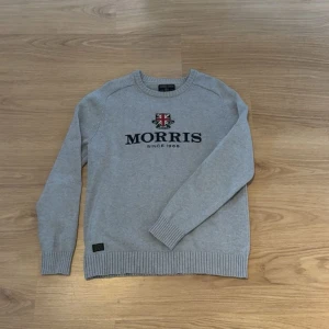 Grå stickad tröja från Morris - Snygg grå stickad tröja från Morris med rund halsringning och broderad logga samt brittisk flagga på bröstet. Tröjan har ribbade muddar vid ärmslut och nederkant. Perfekt för dig som gillar klassisk och stilren design.