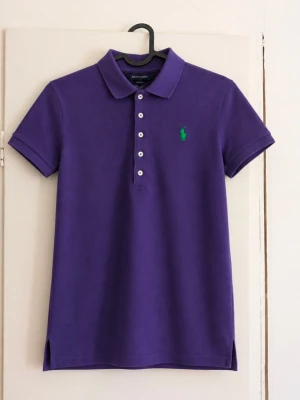 Ralph Lauren lila piké med grön logga – L (12/14) 💜 - Lila pikétröja från Ralph Lauren med grön broderad logga på bröstet. Klassisk modell med krage, fem knappar framtill och små slitsar längst ner. Storlek: L (12/14), barn/ungdom Material: 100% bomull Skick: Mycket fint skick men har nån fläck! Skriv gärna för mått eller fler bilder!