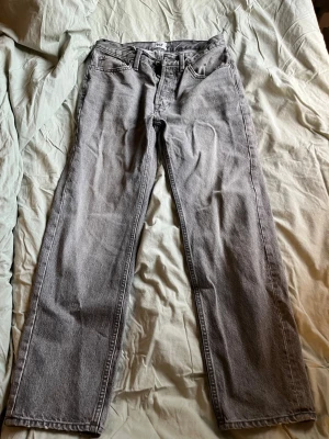 Grå raka jeans med hög midja - Säljer ett par grå jeans med rak passform och hög midja. Klassisk femficksmodell med bälteshällor och dragkedja. Jeansen har en tidlös look och är tillverkade i denim. Perfekta för dig som gillar en clean och enkel stil.