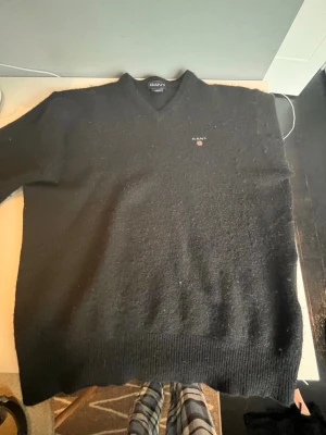 Svart stickad tröja från GANT - Klassisk svart stickad tröja från GANT med V-ringning och broderad logga på bröstet. Tröjan har ribbade muddar vid ärmslut och nederkant. Perfekt för lager på lager och enkel att matcha med olika outfits. Det står att den är Large men den är XXS för att jag har krympt den till min storlek