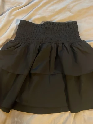 Svart volangkjol med smock - Supersnygg svart kjol med bred smock i midjan och dubbla volanger. Kjolen har en kort modell och är perfekt för dig som gillar en enkel men trendig stil. Materialet känns lätt och flowy, vilket gör den skön att bära. Passar till många olika outfits!