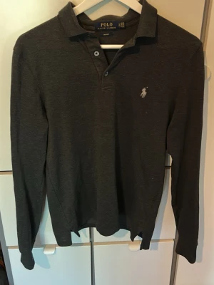 Mörkgrå långärmad piké från Polo Ralph Lauren - Snygg mörkgrå långärmad pikétröja från Polo Ralph Lauren med klassisk krage och två knappar vid halsen. Tröjan har den ikoniska broderade loggan på bröstet och är tillverkad i mjukt bomullsmaterial. Perfekt för en stilren och slimfit look.