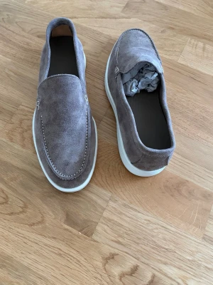 Italienska mockaloafers - Snygga loafers i grå mocka med stilren design och kontrasterande vit sula. Skorna har klassisk slip-on-modell och diskreta sömmar längs ovandelen. Perfekta för dig som gillar en clean och modern look.