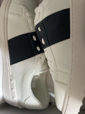Vita sneakers med svart detalj - Snygga vita sneakers med bred svart rem över sidan. Skorna har klassisk låg profil, vita snören och diskreta sömmar. Tillverkade i slätt läderliknande material för en clean look. Perfekta för dig som gillar stilrena och moderna sneakers.