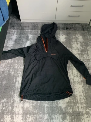 Svart vindjacka från Stormberg - Snygg svart vindjacka från Stormberg med orange dragkedja och detaljer. Jackan har huva, stor magficka och justerbar nederkant med dragsko. Perfekt för blåsiga dagar och outdoor-aktiviteter. Tillverkad i lätt och smidigt material.