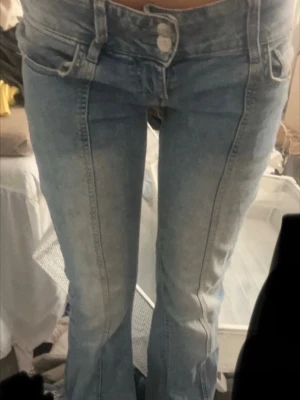 Ljusblå bootcut jeans med låg midja - Säljer ett par ljusblå jeans med bootcut-modell och låg midja. Jeansen har dubbla knappar i linningen, markerade sömmar framtill och klassiska fickor. Perfekta för dig som gillar en retroinspirerad look och vill ha något som sticker ut lite extra.