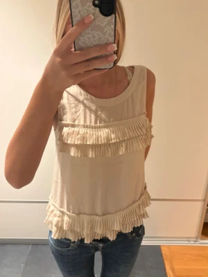 Beige ärmlös topp med volangdetaljer - Säljer en beige ärmlös topp med rund hals och dekorativa volanger framtill och nedtill. Toppen har knappar i ryggen och är gjord i ett lätt, mjukt tyg som känns skönt mot huden. Perfekt att matcha med jeans eller kjol för en trendig look.