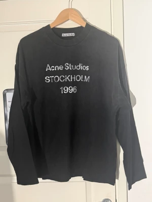 Svart långärmad Acne Studios - Vvart långärmad topp från Acne Studios med tryck i vitt framtill där det står 'Acne Studios STOCKHOLM 1996'. Klassisk rund halsringning och relaxed fit. Perfekt till jeans eller kjol för en clean och trendig look.