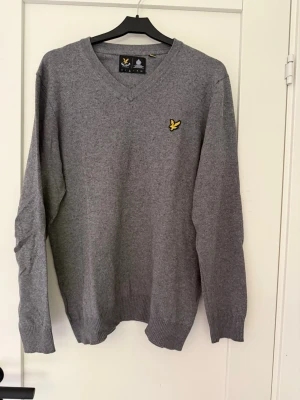 Grå stickad tröja med v-ringning Lyle&Scott - En stilren grå stickad tröja med v-ringad hals och ribbade muddar. Tröjan har en gul broderad logga på bröstet som ger en snygg detalj. Perfekt att matcha med jeans eller chinos för en clean look.