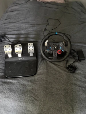 Logitech ratt och pedaler till spelkonsol - Säljer ett Logitech racing set med ratt och pedaler, perfekt för bilspel. Ratten har svart lädergrepp, blå markering och flera knappar, medan pedalerna är i metall och svart plast. Passar till både PC och konsol för en realistisk racingupplevelse.