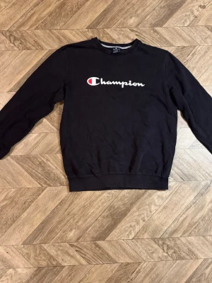 Svart sweatshirt från Champion - Säljer en svart sweatshirt från Champion med klassisk logga i vitt och rött på bröstet. Tröjan har rund halsringning, långa ärmar och ribbade muddar vid ärmslut och nederkant. Perfekt för en avslappnad och sportig stil.