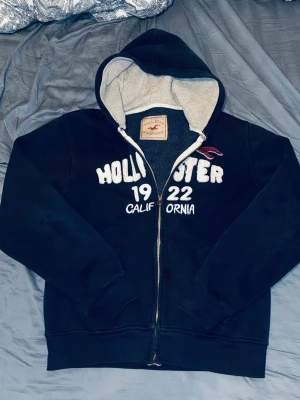 Marinblå zip-hoodie från Hollister - Snygg marinblå hoodie från Hollister med dragkedja, vit broderad text 'Hollister 1922 California' på bröstet och klassisk logga. Mjuk insida i grått tyg och stor huva. Perfekt för en chill och avslappnad stil.